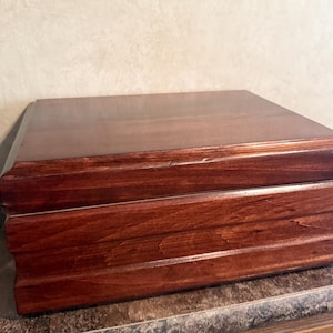 May include: A rectangular wooden box with a rich, dark brown stained finish and a smooth, polished surface. The box features a hinged lid with beveled edges and a tiered, stepped design along the front panel, showcasing natural wood grain patterns.