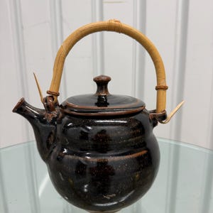 May include: A dark brown glazed ceramic teapot with a speckled finish, featuring a rounded body, a matching lid with a small knob, and a curved spout. The teapot is equipped with a light-colored, arched bamboo handle attached to two loops.