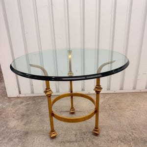 May include: A round side table with a clear glass top and a black rim. The base is constructed from gold-colored metal, featuring three vertical legs connected by a circular support ring, with square-shaped decorative accents at the joints.