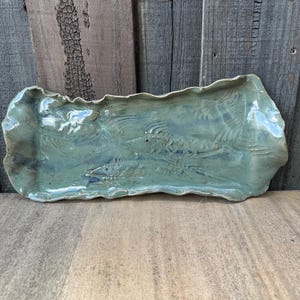 May include: A rectangular ceramic tray with a wavy, uneven rim and a glossy sage green glaze. The surface features an embossed design of two fish swimming in the same direction, with visible scale textures and fins integrated into the clay.