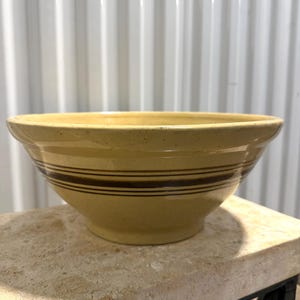 May include: A yellow ceramic mixing bowl with a speckled finish and three horizontal dark brown stripes encircling the exterior. The bowl features a wide, flared rim and a tapered base, showcasing a smooth, glossy glaze throughout the surface.