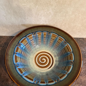 May include: A round ceramic bowl featuring a glossy glaze with a brown rim and a central spiral pattern. The interior displays radiating blue and brown vertical stripes that transition into a cream-colored center with a dark brown spiral design.