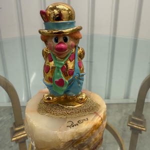 May include: A decorative clown figurine with a gold-colored hat, jacket, and base. The clown has a pink nose, rosy cheeks, and blue overalls. The figurine is mounted on a marble-like base. The signature "Pon ©93" is visible.