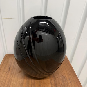 May include: A glossy black ceramic vase with an oval, egg-like shape. The surface features a series of three diagonal, recessed grooves that wrap around the side, creating a textured pattern against the otherwise smooth, reflective finish.