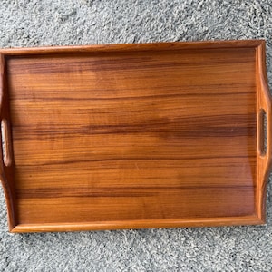May include: A rectangular wooden serving tray featuring a warm, medium-brown wood grain pattern. The tray has raised edges and integrated cutout handles on both the left and right sides, providing a functional design for carrying items.
