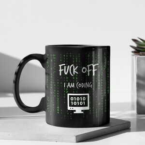 Pode incluir: Caneca de cerâmica preta com o texto branco "FUCK OFF I AM CODING". A caneca apresenta um gráfico de tela de computador com código binário e um fundo em estilo matrix em verde. A caneca tem uma alça preta.