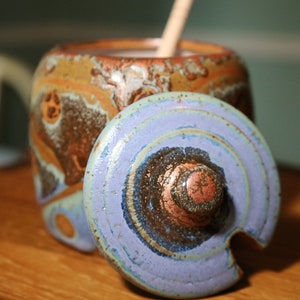 May include: A ceramic honey pot with a blue and brown swirl pattern lid. The pot has a brown and orange swirl pattern.