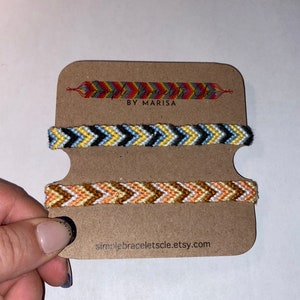 Può includere: Tre braccialetti dell'amicizia fatti a mano con motivi a zig-zag in vari colori sono esposti su una scheda marrone. Il braccialetto superiore presenta un design arcobaleno, quello centrale ha nero, giallo e blu, e quello inferiore è arancione e bianco. La scheda recita "BY MARISA" e "simplebraceletscl.etsy.com".