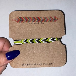 May include: A chevron patterned friendship bracelet in black, white, and neon green, displayed on a brown card. The card features the text "Simple Bracelets Cle" and "by Marisa", along with the Etsy shop URL. The bracelet is handmade.