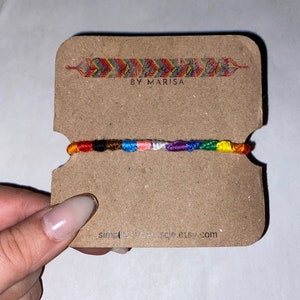 Puede incluir: Una pulsera de la amistad hecha a mano con una combinación de colores del arcoíris, exhibida en una tarjeta marrón. La pulsera presenta una serie de hilos coloridos, incluyendo rojo, naranja, amarillo, verde, azul y morado. La tarjeta tiene el texto "BY MARISA" y "simplemuscle.etsy.com".