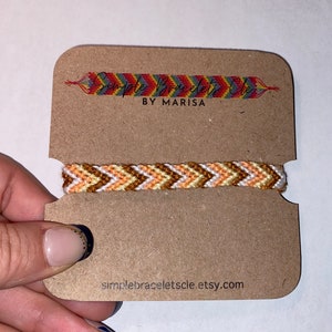 Può includere: Braccialetto dell'amicizia fatto a mano con un motivo a spina di pesce, nei toni dell'arancione, del giallo e del bianco. Il braccialetto è presentato su un cartoncino marrone con il testo "Simple Bracelets by Marisa" e l'URL del negozio Etsy.