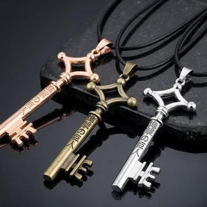 Può includere: Tre collane con ciondoli a forma di chiave in oro rosa, bronzo e argento, ciascuna sospesa a un cordino nero. Le chiavi hanno un design distintivo con una parte superiore a forma di diamante e testo giapponese sullo stelo. I ciondoli sono esposti su una superficie scura.