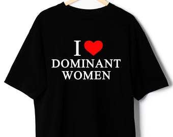 May include: Black t-shirt with the text "I ❤️ DOMINANT WOMEN" in white and a red heart symbol. The shirt is hanging on a wooden hanger against a white background. The t-shirt is a casual wear item.