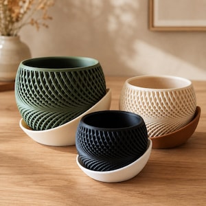 Peut inclure: Trois pots de fleurs ronds avec des motifs en treillis tressés complexes sur l'extérieur. Les pots sont disponibles en vert foncé, crème et noir, chacun reposant sur une soucoupe incurvée assortie ou contrastée, créant un aspect texturé.