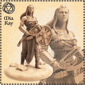 Op de afbeelding: Een miniatuurfiguur van een vrouwelijke krijger met een schild en een bijl. Ze draagt een leren rok en bh. Het figuur is in lichtbruin geverfd.