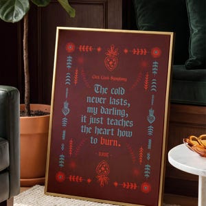 May include: A framed art print with a gold frame. The print has a burgundy background with teal and red decorative elements and text that reads "The cold never lasts, my darling, it just teaches the heart how to burn."