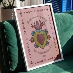 May include: A framed art print with a white border, featuring a pink background and a central illustration of a heart-shaped design with text. The artwork includes celestial and botanical elements, with the quote "The old never lasts, my darling, it just teaches the heart how to burn."