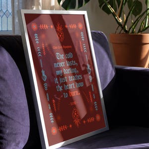 May include: Framed art print with a burgundy background and a white border. The print features decorative floral and heart motifs in orange and teal, with the text "The cold never lasts, my darling, it just teaches the heart how to burn."