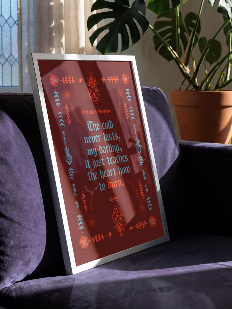 May include: Framed art print with a burgundy background and a white border. The print features decorative floral and heart motifs in orange and teal, with the text "The cold never lasts, my darling, it just teaches the heart how to burn."