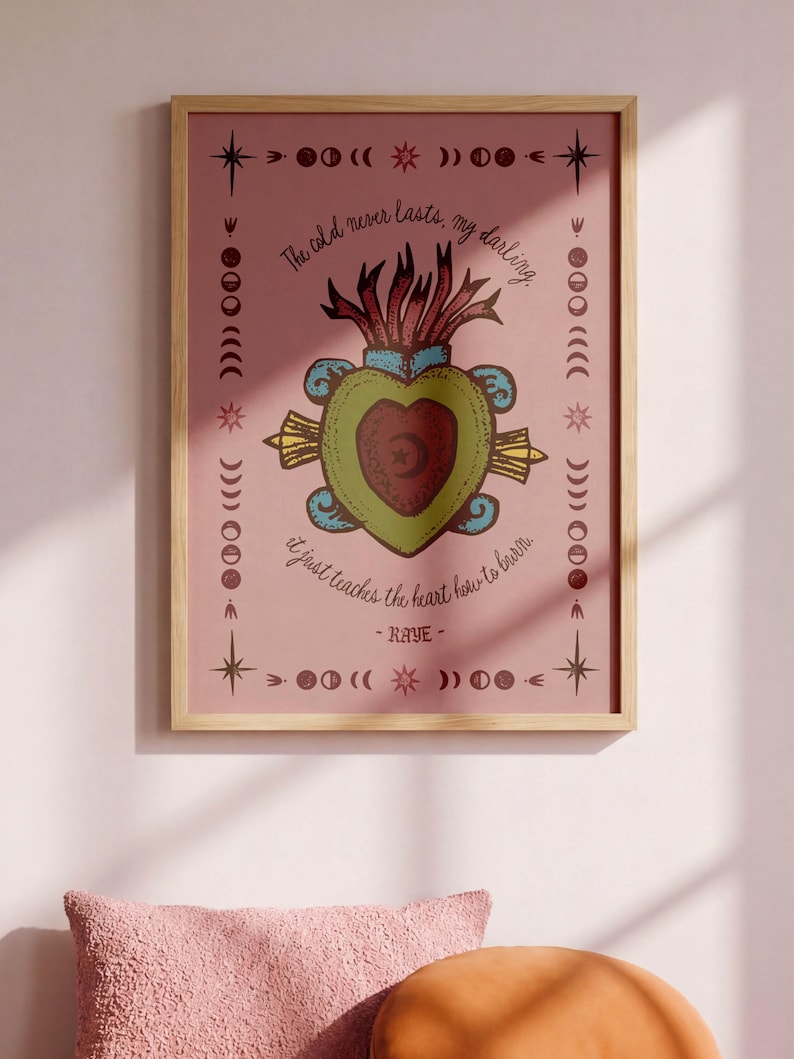 May include: A framed print with a pink background features a heart design with a crescent moon and star. The text reads "The cold never lasts, my darling, it just teaches the heart how to burn." Decorative elements include stars and moon phases.