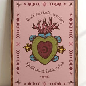 May include: A framed art print with a pink background and a central heart design. The heart is green and maroon, with a crescent moon and star. The print includes the text "The cold never lasts, my darling. It just teaches the heart how to burn. - RAYE -" and decorative celestial elements.