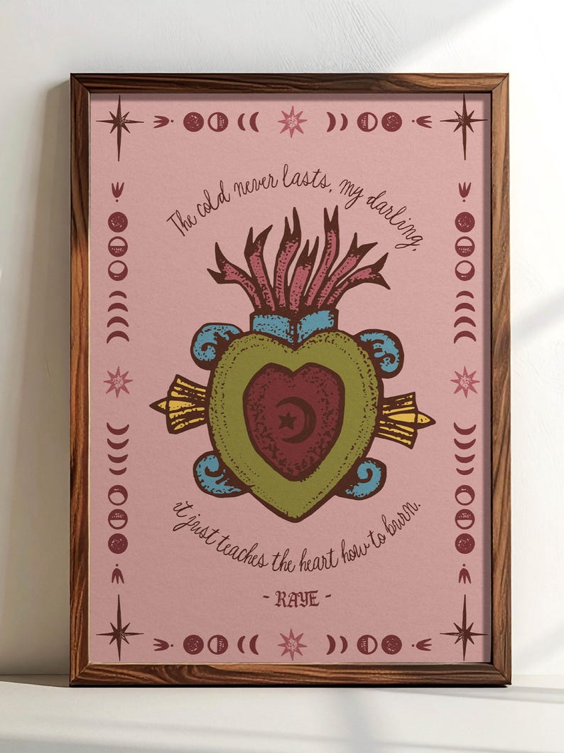 May include: A framed art print with a pink background and a central heart design. The heart is green and maroon, with a crescent moon and star. The print includes the text "The cold never lasts, my darling. It just teaches the heart how to burn. - RAYE -" and decorative celestial elements.