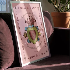 May include: A framed art print with a pink background and a central illustration of a heart-shaped design. The artwork includes text that reads "The cold never lasts, my darling, it just teaches the heart how to burn - RAYE". The frame is white.