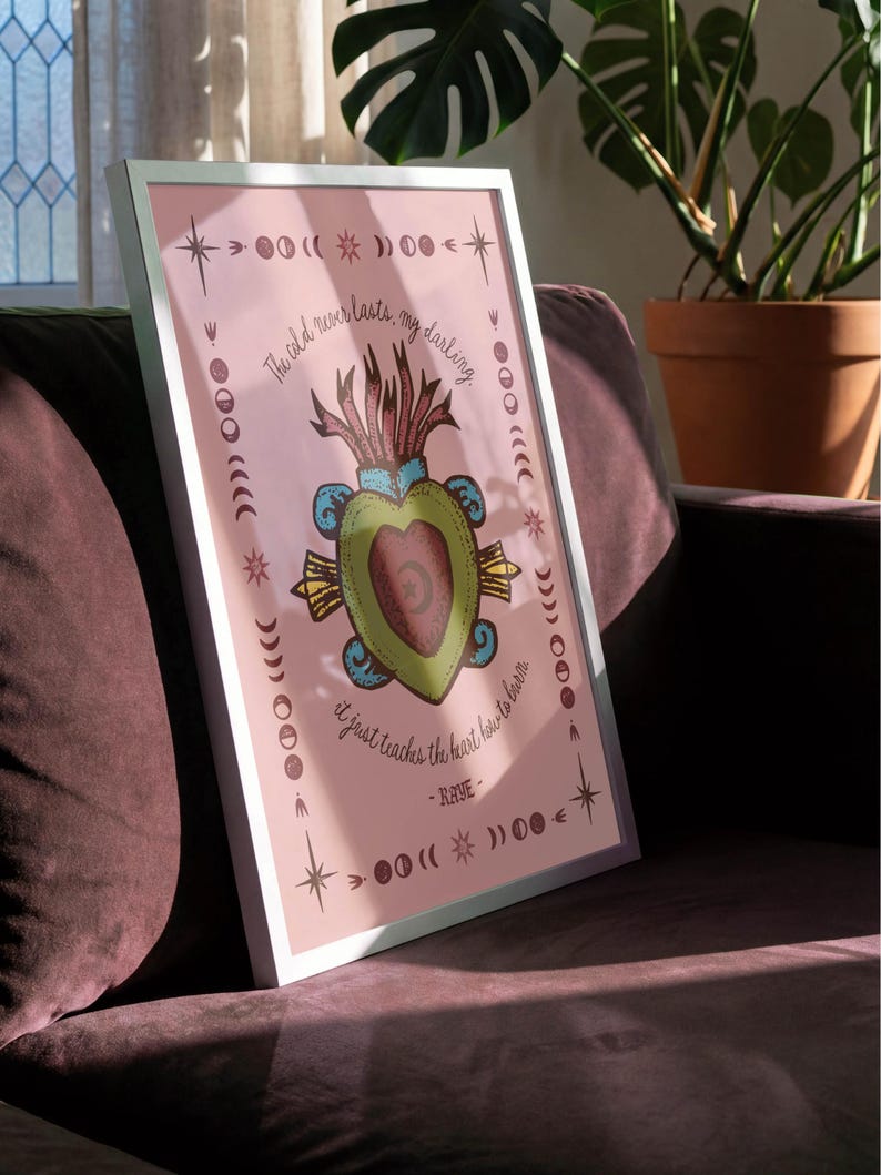 May include: A framed art print with a pink background and a central illustration of a heart-shaped design. The artwork includes text that reads "The cold never lasts, my darling, it just teaches the heart how to burn - RAYE". The frame is white.