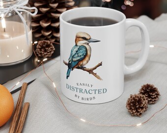 May include: White ceramic mug featuring a watercolor illustration of a bird perched on a branch. The bird has blue, brown, and white feathers. The text "EASILY DISTRACTED BY BIRDS" is printed below the bird in teal.