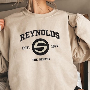 May include: Beige sweatshirt with the text "REYNOLDS EST. 1977 THE SENTRY" in black. The design includes a black circle with a white "S" in the center. The sweatshirt is a neutral color and has a relaxed fit.