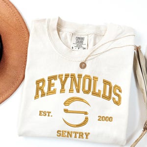May include: Cream-coloured t-shirt with gold embroidered text that reads "REYNOLDS" above a stylized "S" and the words "EST. 2000 SENTRY". A gold necklace with a pendant and a brown hat are also visible.