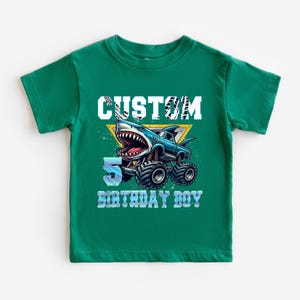 May include: Green short-sleeved t-shirt with a graphic of a blue monster truck with a shark head. The text "CUSTOM 5 BIRTHDAY BOY" is printed on the shirt.