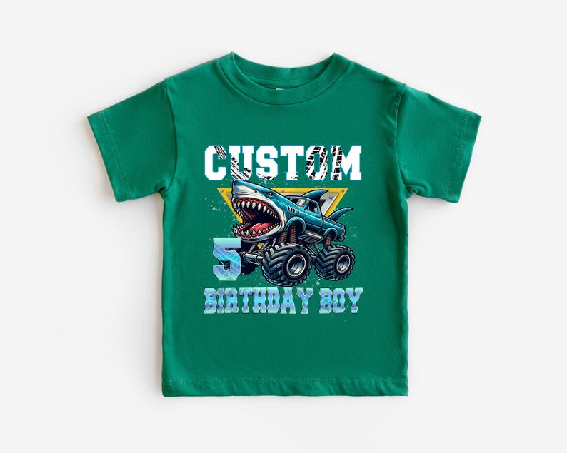 May include: Green short-sleeved t-shirt with a graphic of a blue monster truck with a shark head. The text "CUSTOM 5 BIRTHDAY BOY" is printed on the shirt.