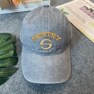 May include: A faded blue baseball cap with the word "SENTRY" embroidered in gold thread across the front. The letter "S" is below the word "SENTRY". The cap has a curved brim and a fabric strap for adjustment.