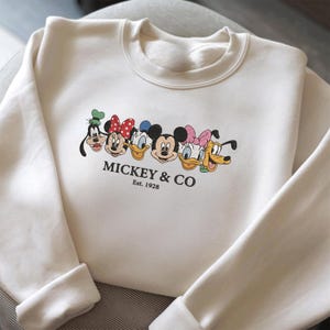 Könnte beinhalten: Cremefarbenes Sweatshirt mit einer Grafik von Mickey Mouse, Minnie Mouse, Donald Duck, Daisy Duck, Goofy und Pluto. Der Text "Mickey & Co. Est. 1928" ist darunter aufgedruckt. Es hat einen Rundhalsausschnitt und lange Ärmel mit gerippten Bündchen.