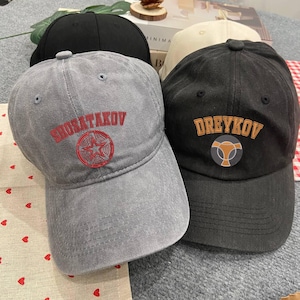 Puede incluir: Dos gorras de béisbol, una gris con bordado rojo que dice "SHORAKOV" con una estrella roja, y una negra con bordado naranja que dice "DREYKOV" con una cabeza de toro estilizada.