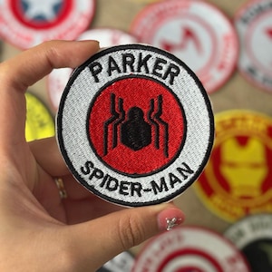 Könnte beinhalten: Gestickter Aufnäher mit dem Text "PARKER SPIDER-MAN" in Weiß auf einem schwarz-weißen kreisförmigen Design. In der Mitte befindet sich ein roter Kreis mit einem schwarzen Spinnensymbol. Weitere Aufnäher sind im Hintergrund zu sehen.