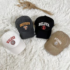 Puede incluir: Cuatro gorras de béisbol con diferentes diseños bordados. Las gorras son de varios colores, incluyendo negro, blanco, gris y marrón. Los diseños incluyen las palabras "Dreykov", "Belova", "Walker" y "Starr", junto con varios símbolos.