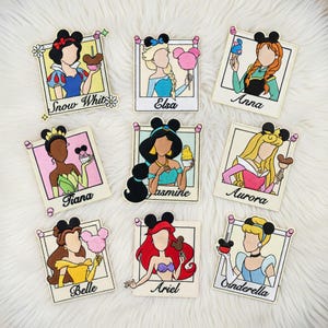 May include: Nine embroidered patches featuring Disney princesses, each in a polaroid-style frame with a Mickey Mouse ear design. The princesses include Snow White, Elsa, Anna, Tiana, Jasmine, Aurora, Belle, Ariel, and Cinderella, each holding a treat.