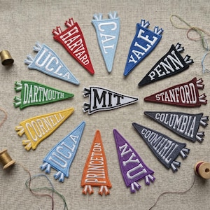Puede incluir: Colección de parches de banderines bordados con nombres de universidades como Harvard, Yale y MIT. Los parches están dispuestos en círculo sobre una superficie de tela neutra, con carretes de hilo y un dedal cerca.