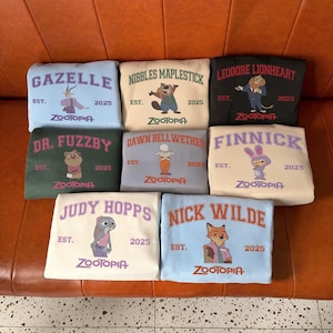 K&ouml;nnte beinhalten: Eine Sammlung von neun Sweatshirts in verschiedenen Farben, die jeweils einen Charakter aus Zootopia zeigen. Die Sweatshirts zeigen Namen wie "Gazelle" und "Judy Hopps" zusammen mit dem Zootopia-Logo und dem Jahr "2025."