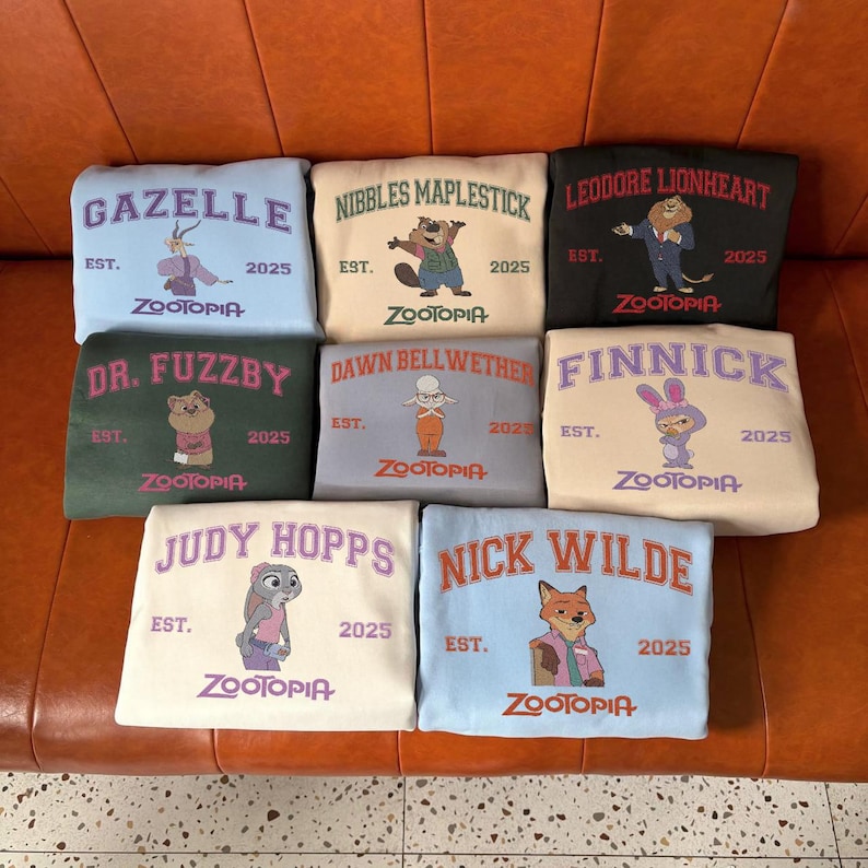 K&ouml;nnte beinhalten: Eine Sammlung von neun Sweatshirts in verschiedenen Farben, die jeweils einen Charakter aus Zootopia zeigen. Die Sweatshirts zeigen Namen wie "Gazelle" und "Judy Hopps" zusammen mit dem Zootopia-Logo und dem Jahr "2025."