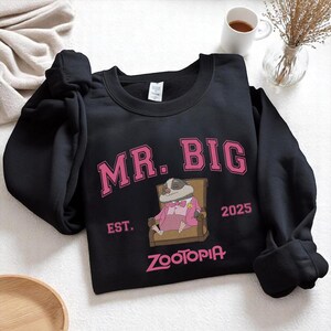 K&ouml;nnte beinhalten: Schwarzes Sweatshirt mit dem pinkfarbenen Schriftzug "MR. BIG" und einer Illustration eines Dachses in einem rosa Anzug. Das Sweatshirt zeigt auch das Wort "ZOOTOPIA" in Pink und das Jahr "2025".