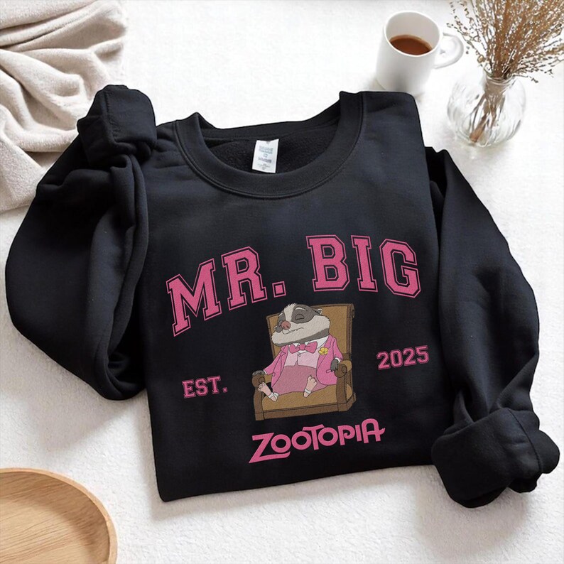 K&ouml;nnte beinhalten: Schwarzes Sweatshirt mit dem pinkfarbenen Schriftzug "MR. BIG" und einer Illustration eines Dachses in einem rosa Anzug. Das Sweatshirt zeigt auch das Wort "ZOOTOPIA" in Pink und das Jahr "2025".