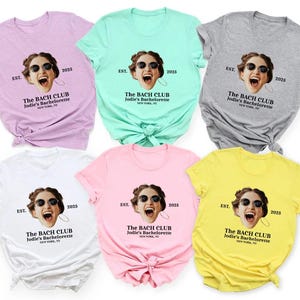 May include: Six different colored t-shirts with a photo of a woman wearing sunglasses and a white shirt with the text "The Bach Club Jodie's Bachelorette EST. 2025" printed on the front.