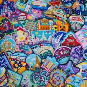 Könnte beinhalten: Eine Sammlung farbenfroher gestickter Aufnäher mit verschiedenen Designs, darunter Burgen, Gebäude und Zeichentrickfiguren. Die Aufnäher weisen leuchtende Farben und detaillierte Stickereien auf, wodurch eine strukturierte und auffällige Darstellung entsteht. Einige Aufnäher haben Text.