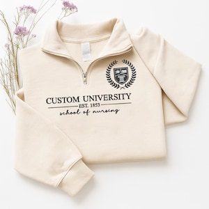 May include: A cream-colored quarter-zip sweatshirt with the text "CUSTOM UNIVERSITY EST. 1853 school of nursing" in black. The sweatshirt has a black emblem of a book and laurel wreath. The garment is folded on a white surface.