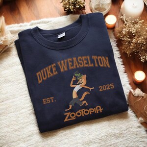 K&ouml;nnte beinhalten: Marineblaues Sweatshirt mit dem Text "DUKE WEASELTON" in brauner, bogenf&ouml;rmiger Schrift und einer Cartoon-Wiesel-Figur mit Hut. Unter der Figur steht das Wort "ZOOTOPIA" und das Jahr "2025". Das Sweatshirt hat einen Rundhalsausschnitt.