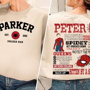 May include: Beige sweatshirt with black text that reads "Parker Est. 2001 Spaider Man" with a red spider graphic. Beige sweatshirt with black text that reads "Peter Parker Friendly Neighborhood Spidey Get Knocked Down Stand Back Up Queens Protect The People 2002 Spider Man On Your Ceiling I Always Get Bitten By A Spider Back Up" with a red spider graphic.
