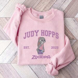 K&ouml;nnte beinhalten: Hellrosa Sweatshirt mit dem Text "JUDY HOPPS" &uuml;ber einer Illustration von Judy Hopps aus Zootopia. Das Sweatshirt enth&auml;lt auch den Text "EST. 2025" und "ZOOTOPIA" in Lila.